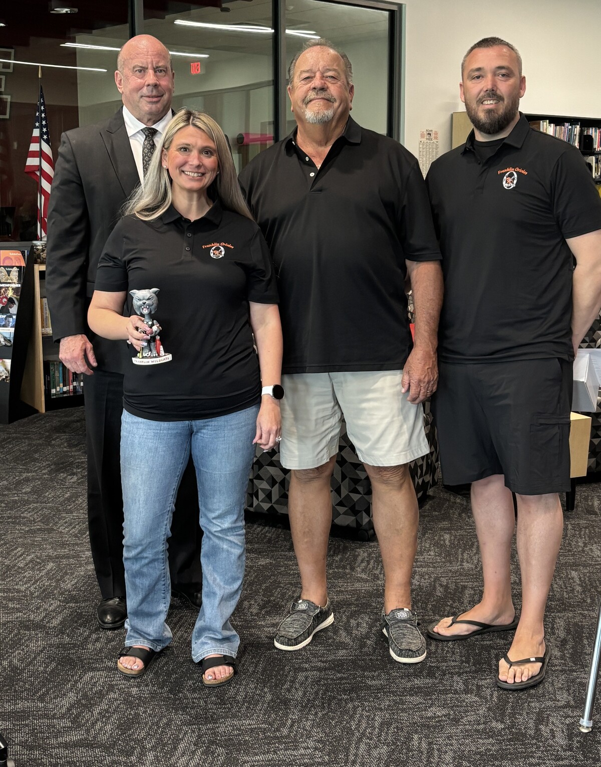 Four individuals in a library with one holding a mascot bobblehead. 